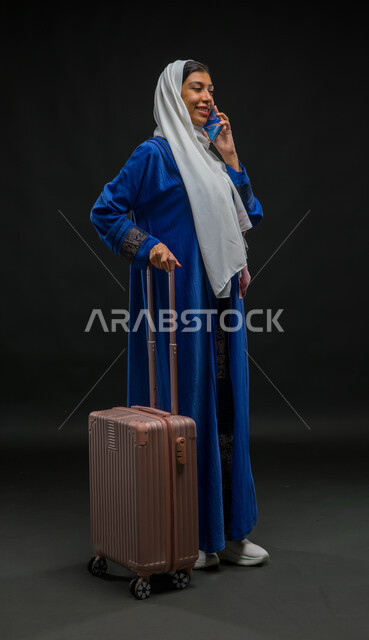 إجراء مكالمة صوتية عبر الهاتف المحمول, التواصل مع الأصدقاء عبر وسائل السوشال ميديا, بورتريه لسيدة عربية خليجية سعودية ترتدي عباءة زرقاء تمسك حقيبة السفر, الاستعداد لبدء الرحلة السياحية بإيماءات البشاشة والسرور, صورة الجسم بالطول الكامل, خلفية سوداء