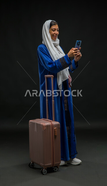 القيام بالبث المباشر والتواصل مع الأصدقاء عبر السوشال ميديا, مفهوم السياحة والسفر, بورتريه لإمرأة عربية خليجية سعودية مبتسمة ترتدي عباءة زرقاء تمسك الهاتف المحمول، استخدام الأجهزة والتقنيات والتطبيقات السياحية, صورة لكامل الجسم , خلفية سوداء