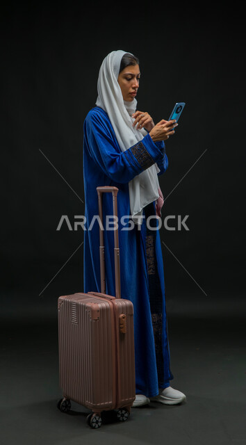 الاستفسار عن شيء ما عبر تطبيقات السياحة والسفر, بورتريه لإمرأة عربية خليجية سعودية ترتدي عباءة زرقاء تمسك الهاتف المحمول بإيماءات الحيرة، استخدام الأجهزة والتقنيات والتطبيقات السياحية, صورة لكامل الجسم , خلفية سوداء