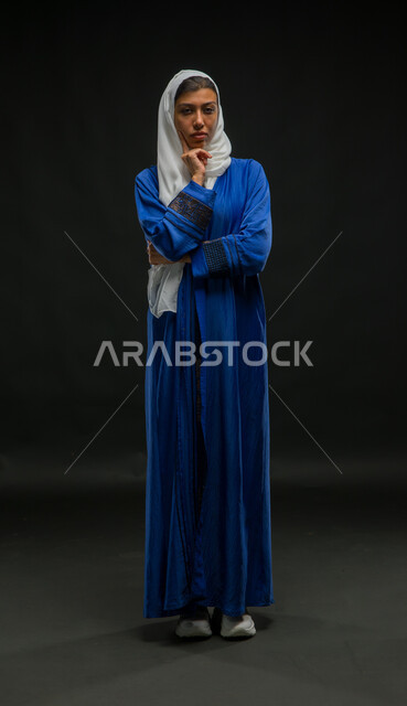 التفكير والتمعن بأمر ما، إيجاد الحل لمشكلة ما، بورتريه لإمرأة عربية سعودية خليجية محجبة ترتدي العباءة الزرقاء تضع يدها تحت رأسها بإيماءات الحيرة والتخطيط للمستقبل، الشعور بالتعب والإرهاق، صورة الجسم بالطول الكامل، خلفية سوداء