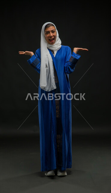 الذهول والمفاجأة من شيء ما، مفهوم النعومة والانوثة والاناقة، بورتريه لإمرأة عربية سعودية خليجية مبتسمة محجبة ترتدي العباءة الزرقاء تقف باستقامة ترفع يديها نحو الأعلى بإيماءات الدهشة، صورة الجسم بالطول الكامل، خلفية سوداء