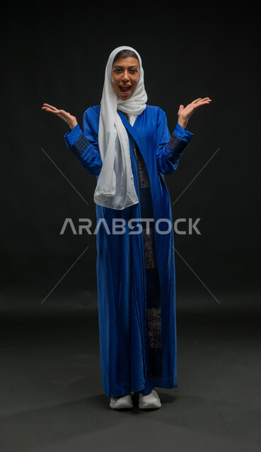 Standing straight and looking at the camera with surprised gestures, the concept of softness, femininity and elegance, a portrait of a smiling Saudi Arabian Gulf woman, veiled, wearing a blue abaya, raising her hands upward with an expression of astonishment, full-length body portrait, black backgroun