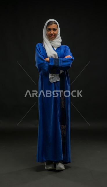 The concept of softness, femininity and elegance, gestures of self-confidence, attention to external appearance, a portrait of a smiling Saudi Arabian Gulf woman, veiled and wearing a blue abaya, standing straight and looking at a camera with crossed hand movements, full-length body portrait, black background