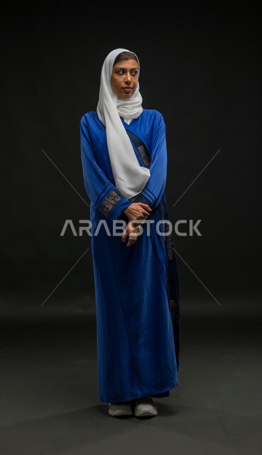 Concern with external appearance, the concept of femininity and elegance, a portrait of a smiling, veiled Saudi Arabian Gulf woman wearing a blue abaya, standing straight and looking to the side with coherent hand movements, full-length body portrait, black background