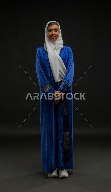 مفهوم الانوثة والاناقة، الاهتمام بالمظهر الخارجي، بورتريه لإمرأة عربية سعودية خليجية مبتسمة محجبة ترتدي العباءة الزرقاء تقف باستقامة وتنظر إلى الكاميرا بحركات أيدي متماسكة، صورة الجسم بالطول الكامل، خلفية سوداء