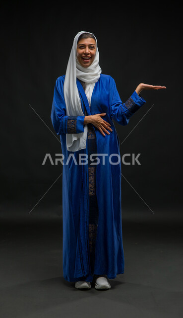 النظر إلى الكاميرا بإيماءات البشاشة والإعجاب، ارتداء أزياء محجبات عصرية، مفهوم الأنوثة والنعومة، بورتريه لامرأة عربية خليجية سعودية محجبة ترتدي عباءة زرقاء ترفع يدها وتحمل عنصر ما بتعابير السرور والذهول، صورة لكامل الجسم، خلفية سوداء