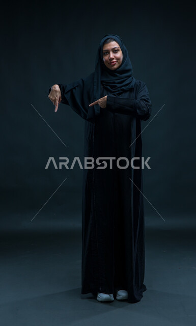 إشارات وحركات يدين تدل على هذا المكان (هنا)، تحديد موقع معين بإيماءات السرور، بورتريه لإمرأة عربية سعودية خليجية ترتدي الحجاب والعباءة التقليدية تشير بأصابعها نحو الأسفل، صورة الجسم بالطول الكامل، خلفية سوداء