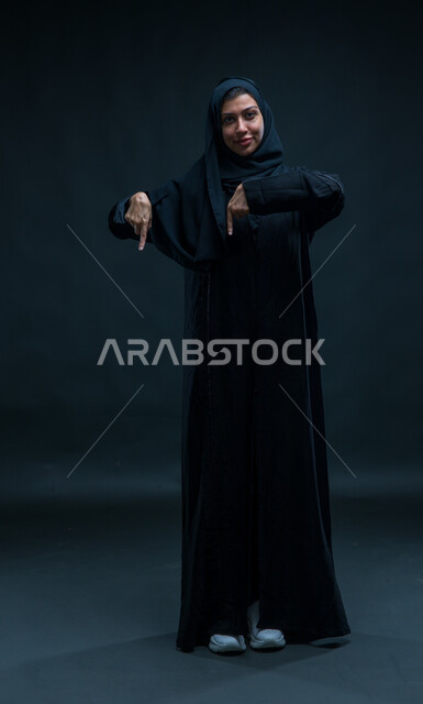 إشارات وحركات يدين تدل على هذا المكان (هنا)، تحديد موقع معين بإيماءات السرور، بورتريه لإمرأة عربية سعودية خليجية ترتدي الحجاب والعباءة التقليدية تشير بأصابعها نحو الأسفل، صورة الجسم بالطول الكامل، خلفية سوداء