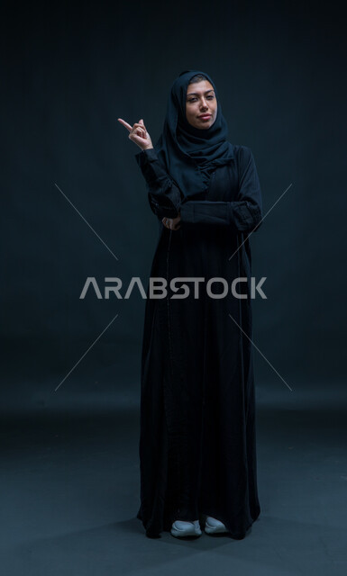 تعابير التحديد والتأكيد, الاشارة إلى شيء ما, بورتريه لسيدة عربية خليجية سعودية ترتدي الحجاب والعباءة التقليدية تشير بأصبعها السبابة لاختيار شيء معين وتنظر إلى الكاميرا، صورة الجسم بالطول الكامل، خلفية سوداء