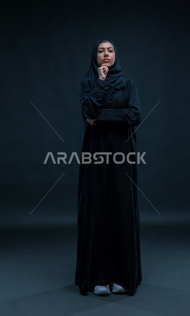 محاولة إيجاد حل لمشكلة ما, التمعن والتركيز والتخطيط للمستقبل، بورتريه لإمرأة عربية خليجية سعودية ترتدي الحجاب والعباءة التقليدية تضع يدها على ذقنها وتنظر إلى الكاميرا بإيماءات التفكير والتأمل، صورة الجسم بالطول الكامل، خلفية سوداء