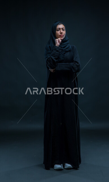 التمعن والتركيز والتخطيط للمستقبل، محاولة إيجاد حل لمشكلة ما، بورتريه لإمرأة عربية خليجية سعودية ترتدي الحجاب والعباءة التقليدية تضع يدها على ذقنها وتنظر إلى الكاميرا بإيماءات التفكير والتأمل، صورة الجسم بالطول الكامل، خلفية سوداء