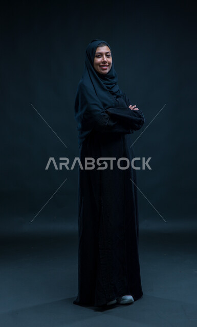 Looking at the camera with crossed hand gestures, the concept of elegance and concern for external appearance, standing upright and self-confident, portrait of a Saudi Arabian Gulf woman wearing the hijab and traditional abaya, standing upright, full-length body portrait, black background