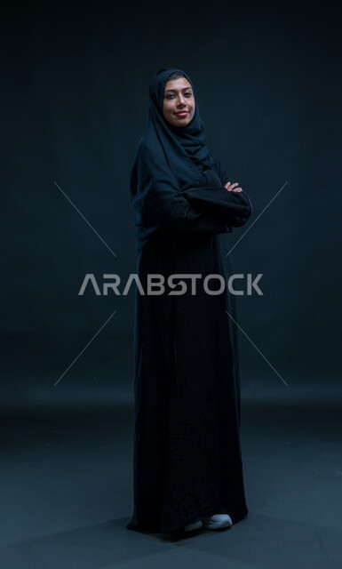 الوقوف باستقامة وثقه بالنفس, مفهوم الاناقه و الاهتمام بالمظهر الخارجي، ، بورتريه لإمرأة عربية خليجية سعودية ترتدي الحجاب والعباءة التقليدية تقف باستقامة، النظر إلى الكاميرا بإيماءات أيدي متقاطعه، صورة الجسم بالطول الكامل، خلفية سوداء