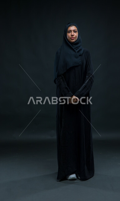 النظر إلى الكاميرا بإيماءات الجدية، مفهوم الاناقه و الاهتمام بالمظهر الخارجي، الوقوف باستقامة وثقه بالنفس، بورتريه لإمرأة عربية خليجية سعودية ترتدي الحجاب والعباءة التقليدية تقف باستقامة بأيدي متماسكة، صورة الجسم بالطول الكامل، خلفية سوداء