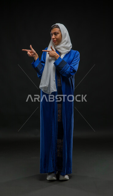 تحديد موقع وشيء معين، إشارات وحركات يدين تدل على هذا المكان (هنا)، بورتريه لامرأة عربية خليجية سعودية محجبة ترتدي عباءة زرقاء تشير بإصبع السبابة إلى جهة اليمين، صورة الجسم بالكامل، خلفية سوداء