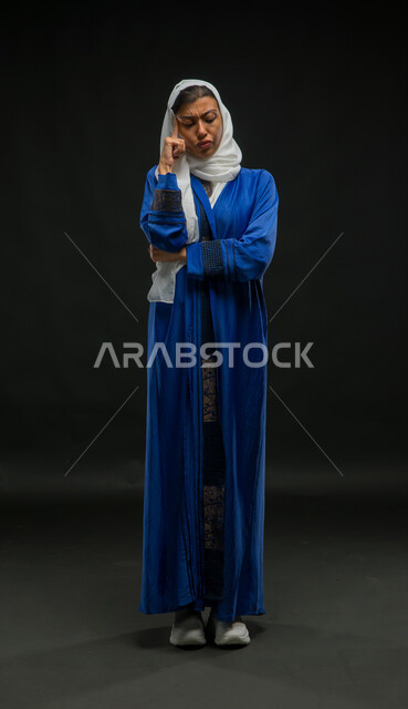Feeling upset and bored, contemplating and focusing to find the solution to a problem, solving problems and making decisions, portrait of a veiled Saudi Gulf Arab woman wearing a blue abaya, placing her hand on her face with gestures of thought, confusion and annoyance, full body portrait, black background