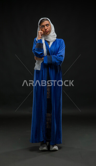 حل المشكلات واتخاذ القرارات، النظر إلى الأعلى بتعابير التخطيط لإيجاد الحل لمشكلة ما, بورتريه لامرأة عربية خليجية سعودية محجبة ترتدي عباءة زرقاء تضع يدها على وجهها بإيماءات التفكير والحيرة، صورة الجسم بالكامل، خلفية سوداء