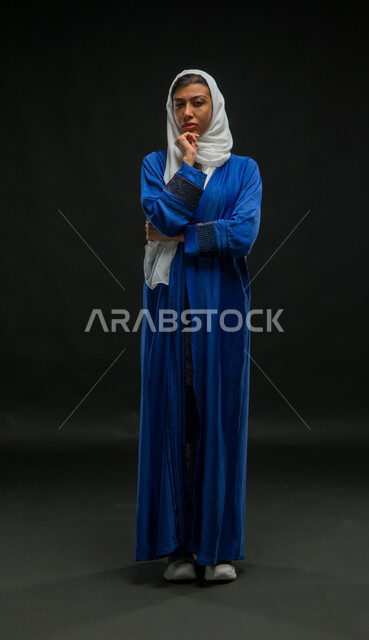 التفكر والتخطيط لإيجاد الحل لمشكلة ما, حل المشكلات واتخاذ القرارات، بورتريه لامرأة عربية خليجية سعودية محجبة ترتدي عباءة زرقاء تضع يدها على وجهها بإيماءات التفكير والحيرة، صورة الجسم بالكامل، خلفية سوداء