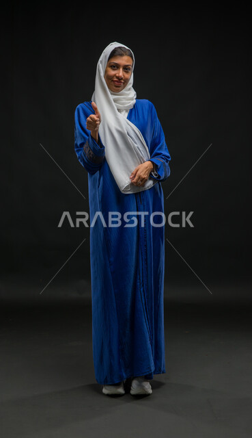 النظر إلى الكاميرا بإيماءات السرور والإعجاب، ارتداء أزياء محجبات عصرية، مفهوم الأنوثة والنعومة، بورتريه لامرأة عربية خليجية سعودية محجبة ترتدي عباءة زرقاء تشير بعلامة الجودة والرضا والامتياز، صورة لكامل الجسم، خلفية سوداء