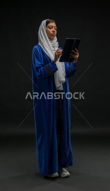 Recording and writing data and notes, using modern technical devices, a portrait of a veiled Saudi Gulf Arab woman wearing a blue abaya performing work tasks via a tablet, expressions and gestures of focus and integration, the concept of remote project management, full body image, black background