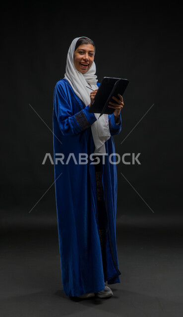 النظر الى الكاميرا بتعابير الفرح والسرور, تدوين البيانات والملاحظات، إستخدام اجهزة تقنية حديثة، بورتريه لإمرأة عربية خليجية سعودية محجبة ترتدي عباءة زرقاء تقوم بانجاز مهام العمل عبر جهاز التابلت, مفهوم إدارة المشاريع عن بعد, صورة الجسم بالكامل، خلفية سوداء