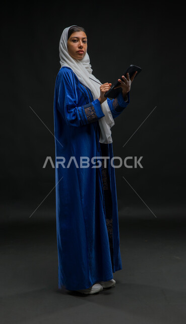 النظر الى الكاميرا بإيماءات الجدية والثقة, تدوين البيانات والملاحظات، إستخدام اجهزة تقنية حديثة، بورتريه لإمرأة عربية خليجية سعودية محجبة ترتدي عباءة زرقاء تقوم بانجاز مهام العمل عبر جهاز التابلت, مفهوم إدارة المشاريع عن بعد, صورة الجسم بالكامل، خلفية سوداء
