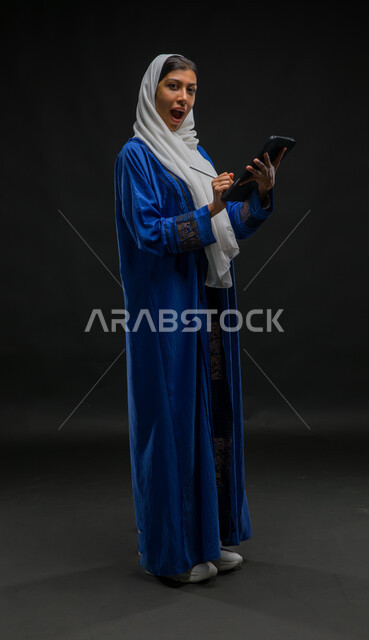 Looking at the camera with expressions of enthusiasm, writing down and recording data and notes, using modern technical devices, a portrait of a veiled Saudi Gulf Arab woman wearing a blue abaya, completing work tasks via a tablet, the concept of remote project management, full body image, black background