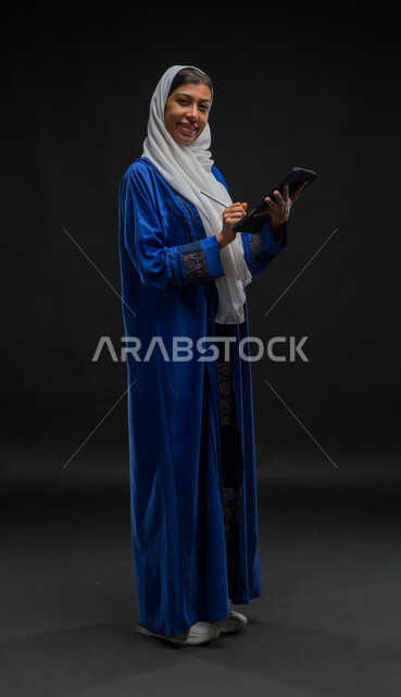 The concept of managing projects remotely, looking at the camera with happy expressions, writing down data and notes, using modern technical devices, a portrait of a smiling Saudi Gulf Arab woman wearing a hijab and a blue abaya, completing work tasks via a tablet, full body portrait, black background