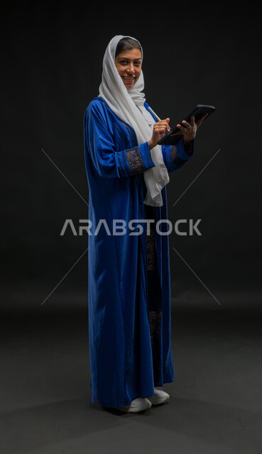 Completing work tasks via a tablet, the concept of remote project management, looking at the camera with happy expressions, using modern technical devices, a portrait of a smiling Saudi Arabian Gulf woman wearing a hijab and a blue abaya, writing down data and notes, full body photo, black background
