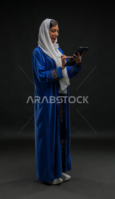 إيماءات وتعابير الحماس والسعادة, تدوين وتسجيل البيانات والملاحظات، إستخدام اجهزة تقنية حديثة، بورتريه لإمرأة عربية خليجية سعودية محجبة ترتدي عباءة زرقاء تقوم بانجاز مهام العمل عبر جهاز التابلت, مفهوم إدارة المشاريع عن بعد, صورة الجسم بالكامل، خلفية سوداء