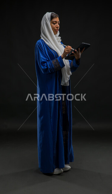Integration into recording and writing data and notes, using modern technical devices, portrait of a veiled Saudi Arabian Gulf woman wearing a blue abaya performing work tasks via a tablet, concentration expressions and gestures, the concept of remote project management, full body image, black background