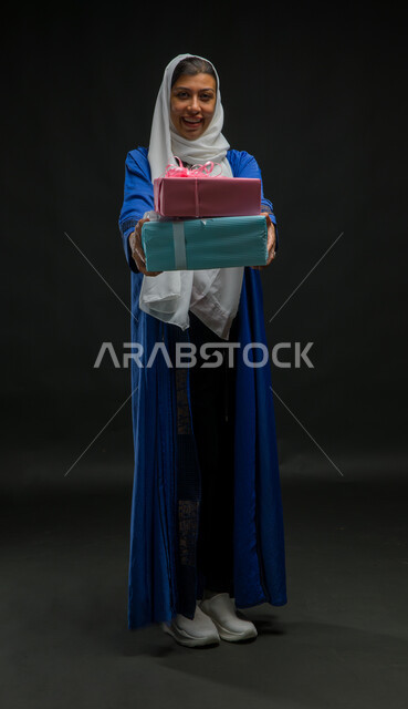 Happiness in giving gifts and congratulations, holidays, occasions and happy surprises, facial gestures indicating shyness and pleasure, portrait of a smiling Saudi Arabian Gulf woman wearing a hijab and blue abaya, holding wrapped gift boxes in her hands, full-length body portrait, black background