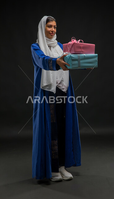 Giving gifts and congratulations, holidays, occasions and happy surprises, facial gestures indicating shyness and pleasure, portrait of a smiling Saudi Arabian Gulf woman wearing a hijab and blue abaya, holding wrapped gift boxes in her hands, full-length body portrait, black background