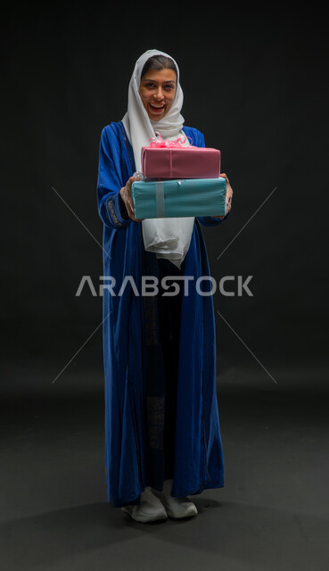 Looking at the camera with facial gestures indicating astonishment and pleasure, the joy of receiving gifts and congratulations, a portrait of a veiled Saudi Arabian Gulf woman wearing a blue abaya, holding wrapped gift boxes in her hands, full-length body portrait, holidays, occasions and happy surprises, black background