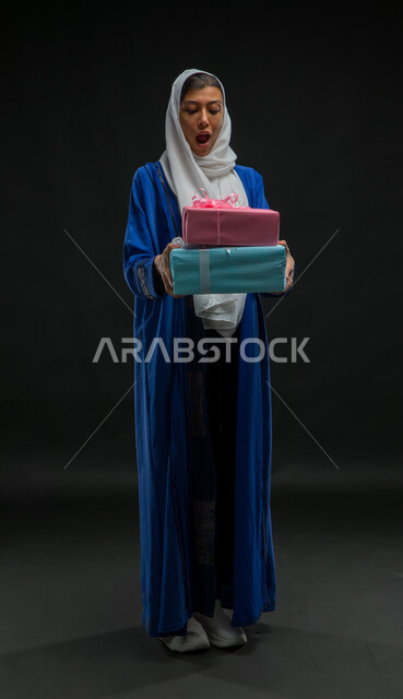 أعياد ومناسبات ومفاجآت سعيدة, إيماءات وجه تدل على الاندهاش والفرح بالحصول على الهدايا والتهنئات، بورتريه لامرأة عربية سعودية خليجية محجبة ترتدي عباءة زرقاء تحمل بيديها علب هدايا مغلفة، صورة الجسم بالطول الكامل، خلفية سوداء