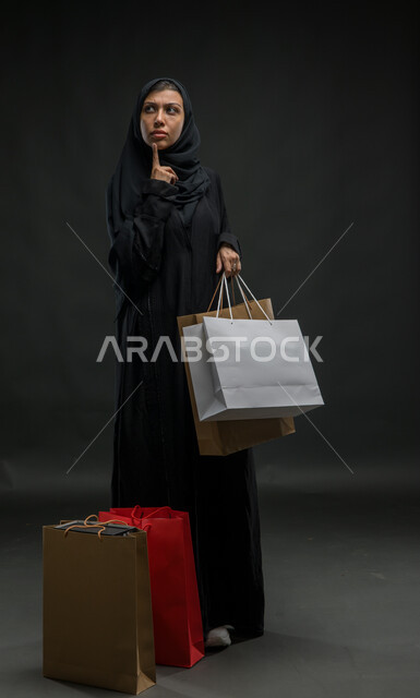 تعابير التمعن والتركيز، إيماءات التفكير والتخطيط، الاستفادة من الخصومات والتخفيضات، بورتريه لإمرأة عربية خليجية سعودية محجبة ترتدي العباءة السوداء تحمل بيدها أكياس تسوق ملونة، شراء المستلزمات والهدايا، صورة الجسم بالكامل، خلفية سوداء