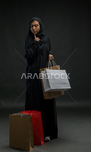إيماءات التفكير والتخطيط، الاستفادة من الخصومات والتخفيضات، بورتريه لإمرأة عربية خليجية سعودية محجبة ترتدي العباءة السوداء تحمل بيدها أكياس تسوق ملونة، تعابير التمعن والتركيز، شراء المستلزمات والهدايا، صورة الجسم بالكامل، خلفية سوداء