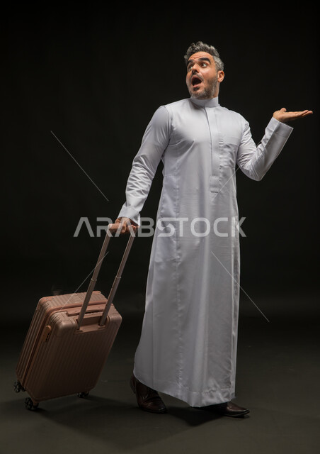 تعابير الذهول والتفاجؤ والاستغراب, بورتريه لرجل عربي خليجي سعودي يرتدي الثوب التقليدي يمسك حقيبة السفر ويرفع كفه للأعلى, النظر إلى شيء ما بإيماءات الدهشة, محاولة رفع عنصر ما, صورة الجسم بالطول الكامل, خلفية سوداء