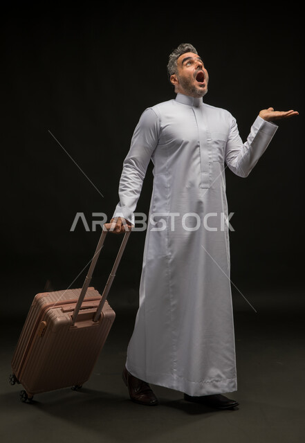 Expressions of astonishment, surprise, and astonishment, portrait of a Saudi Gulf Arab man wearing a traditional thobe holding a suitcase and raising his palm up, looking at something with gestures of astonishment, trying to lift an item, full-length body portrait, black background