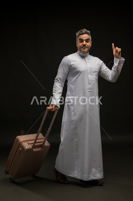النظر إلى الكاميرا والإشارة إلى شيء ما, السياحة في إجازة الصيف, بورتريه لرجل عربي سعودي خليجي يرتدي الثوب التقليدي يقوم بجر حقيبة السفر ويشير بإصبعه السبابة للأعلى, الاستعداد لقضاء رحلة صيفية ممتعة, صورة الجسم بالطول الكامل, خلفية سوداء