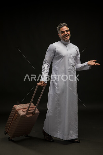 الوقوف باستقامة والإشارة إلى مكان معين, بورتريه لرجل عربي خليجي سعودي مبتسم يرتدي الثوب التقليدي يمسك حقيبة السفر ويفتح كفه بإيماءات تحديد شيء ما ببشاشة وسرور, مفهوم السياحة والوجهات السياحية الترفيهيه, صورة الجسم بالطول الكامل, خلفية سوداء