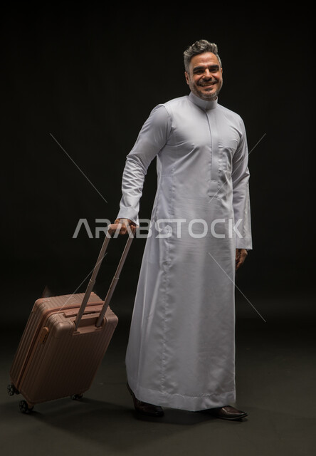 الوقوف باستقامة وانتظار موعد الطائرة, مفهوم السياحة والقيام بالجولات الترفيهية في وقت الإجازة, بورتريه لرجل عربي خليجي سعودي مبتسم يرتدي الثوب التقليدي يقوم بجر حقيبة السفر, الاستعداد لبدء الرحلة في العطلة الصيفيه بسرور, صورة الجسم بالطول الكامل, خلفية سوداء