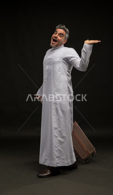 تعابير الذهول والتفاجؤ والاستغراب, بورتريه لرجل عربي خليجي سعودي يرتدي الثوب التقليدي يمسك حقيبة السفر ويرفع كفه للأعلى, النظر إلى الكاميرا بإيماءات الدهشة, محاولة رفع عنصر ما, صورة الجسم بالطول الكامل, خلفية سوداء