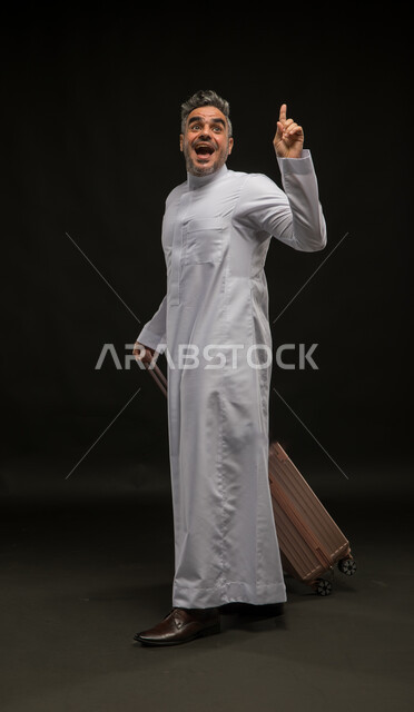 النظر إلى الأعلى بإيماءات الذهول والإشارة إلى شيء ما, السياحة في إجازة الصيف, بورتريه لرجل عربي سعودي خليجي يرتدي الثوب التقليدي يقوم بجر حقيبة السفر, الاستعداد لقضاء رحلة صيفية ممتعة, الشعور بالحماس والفرح، صورة الجسم بالطول الكامل, خلفية سوداء