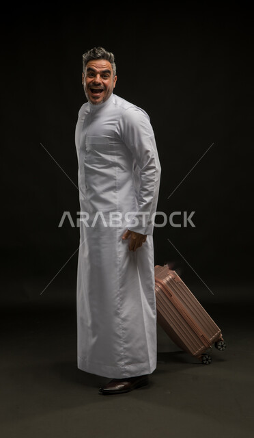 الذهول والاستغراب والتفاجؤ,السرور والبشاشة لرؤية شخص ما, بورتريه لرجل عربي خليجي سعودي يرتدي الثوب التقليدي يمسك حقيبة السفر بإيماءات الدهشة والسعادة, الإعجاب بمكان معين, الاستمتاع بالرحلة السياحية برفقة الأصدقاء, صورة الجسم بالطول الكامل, خلفية سوداء