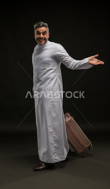 الوقوف بذهول والإشارة إلى مكان معين, بورتريه لرجل عربي خليجي سعودي مبتسم يرتدي الثوب التقليدي يمسك حقيبة السفر ويفتح كفه بإيماءات تحديد شيء ما ببشاشة وسرور, مفهوم السياحة والوجهات السياحية الترفيهيه, صورة الجسم بالطول الكامل, خلفية سوداء