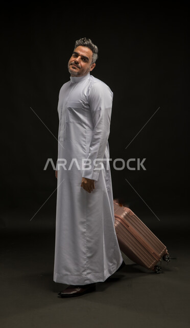 Looking at the camera with gestures of dissatisfaction and sadness, plane delay, portrait of a Saudi Gulf Arab man wearing a traditional dress dragging a suitcase, concept of tourism and leisure trips, full-length body portrait, black background