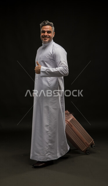 الاستعداد لبدء الرحلة الصيفيه بسرور, مفهوم السياحة والقيام بالجولات الترفيهية بإيماءات الجودة والامتياز والرضى, الوقوف باستقامة وانتظار موعد الطائرة, بورتريه لرجل عربي خليجي سعودي مبتسم يرتدي الثوب التقليدي يقوم بجر حقيبة السفر, صورة الجسم بالكامل, خلفية سوداء