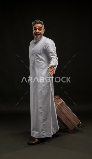 السرور والبشاشة لرؤية شخص ما, بورتريه لرجل عربي خليجي سعودي يرتدي الثوب التقليدي يمسك حقيبة السفر بإيماءات الدهشة والسعادة, الإعجاب بمكان معين, الاستمتاع بالرحلة السياحية برفقة الأصدقاء, صورة الجسم بالطول الكامل, خلفية سوداء