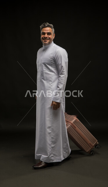 The concept of tourism and taking recreational tours during vacation time, standing straight and waiting for the plane to arrive, portrait of a smiling Saudi Gulf Arab man wearing a traditional dress dragging a travel bag, preparing to start the trip on the summer vacation with pleasure, full-length body portrait, black background
